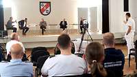 Fünf Personen sitzen auf einer Bühne vor Publikum. Ein Mann aus dem Plenum spricht zu den Personen auf der Bühne.
