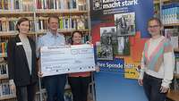 Drei Frauen und ein Soldat stehen in einer Bibliothek und präsentieren einen Spendenscheck