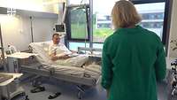 Eine Frau in grünem Oberteil geht auf einen Mann im Krankenbett zu