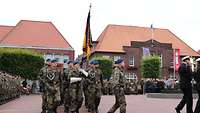 Mehrere Soldaten marschieren in Formation auf einen rot gepflasterten Platz. In der Mitte ist eine Fahne zu sehen.