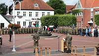 Auf einen Marktplatz stehen viele angetretene Soldaten. In Front vor ihnen steht ein Soldat am Rednerpult
