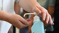 Ausschnitt von zwei Händen. Dabei drückt eine Hand auf eine Desinfektionsflasche und die andere ist unter der Öffnung