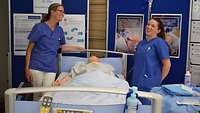 Zwei Frauen in Krankenhauskleidung stehen an einem Patientenbett.