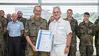 Zwei Soldaten halten eine Zertifikatsurkunde in der Hand. Im Hintergrund stehen weitere Soldaten.