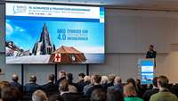 Ein Oberstarzt steht am Pult und begrüßt Menschen auf einem Kongress