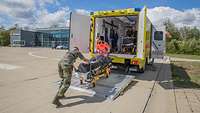Eine militärisch und eine zivil gekleidete Rettungskraft bewegt eine Trage auf die Hebebühne des Intensivtransportwagens