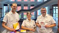 Eine Frau und zwei Männer vor einem Tisch mit Broschüren.